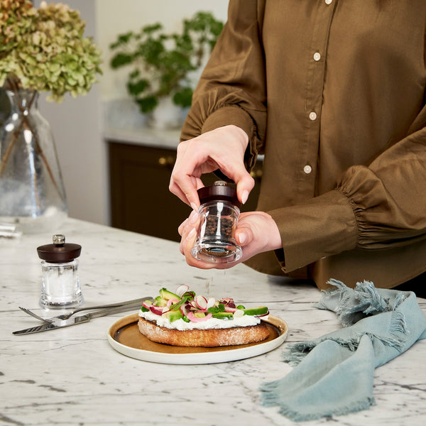 A person in a brown shirt uses a pepper grinder to season an open-faced sandwich with vegetables on a plate, on a marble kitchen counter with a napkin, fork, and knife nearby.