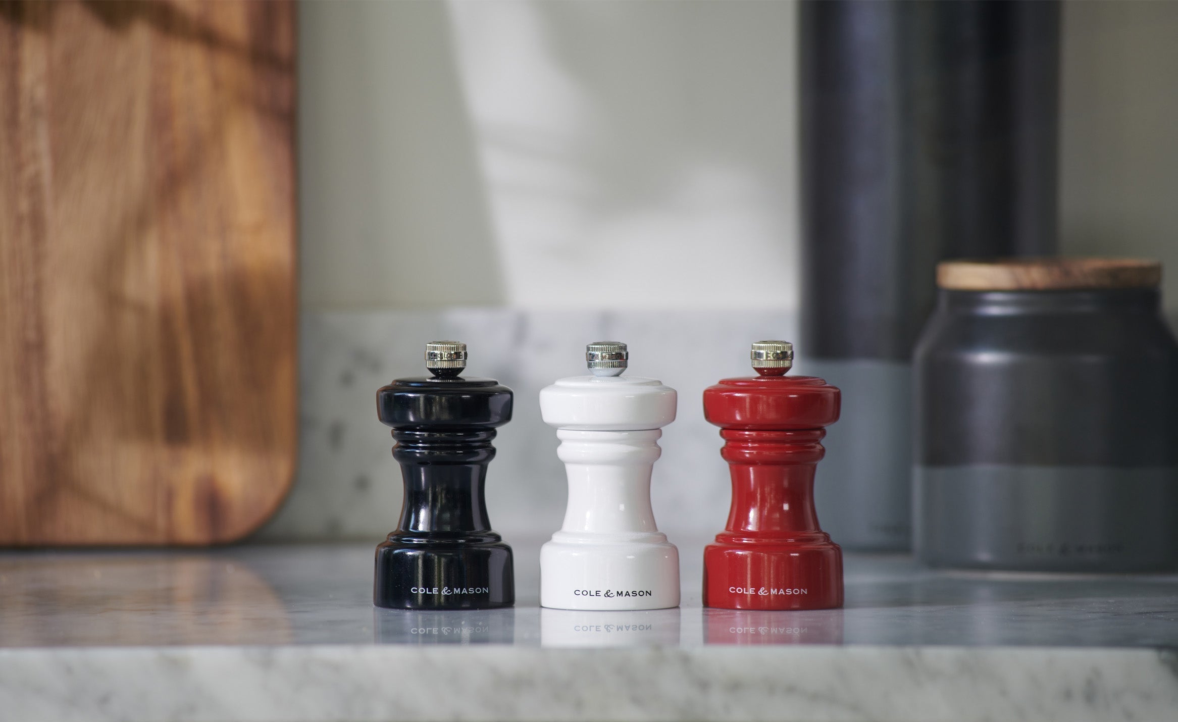 Three Cole & Mason spice grinders—black, white, and red—stand in a row on a marble countertop. A wooden board and dark grey container are blurred in the background.