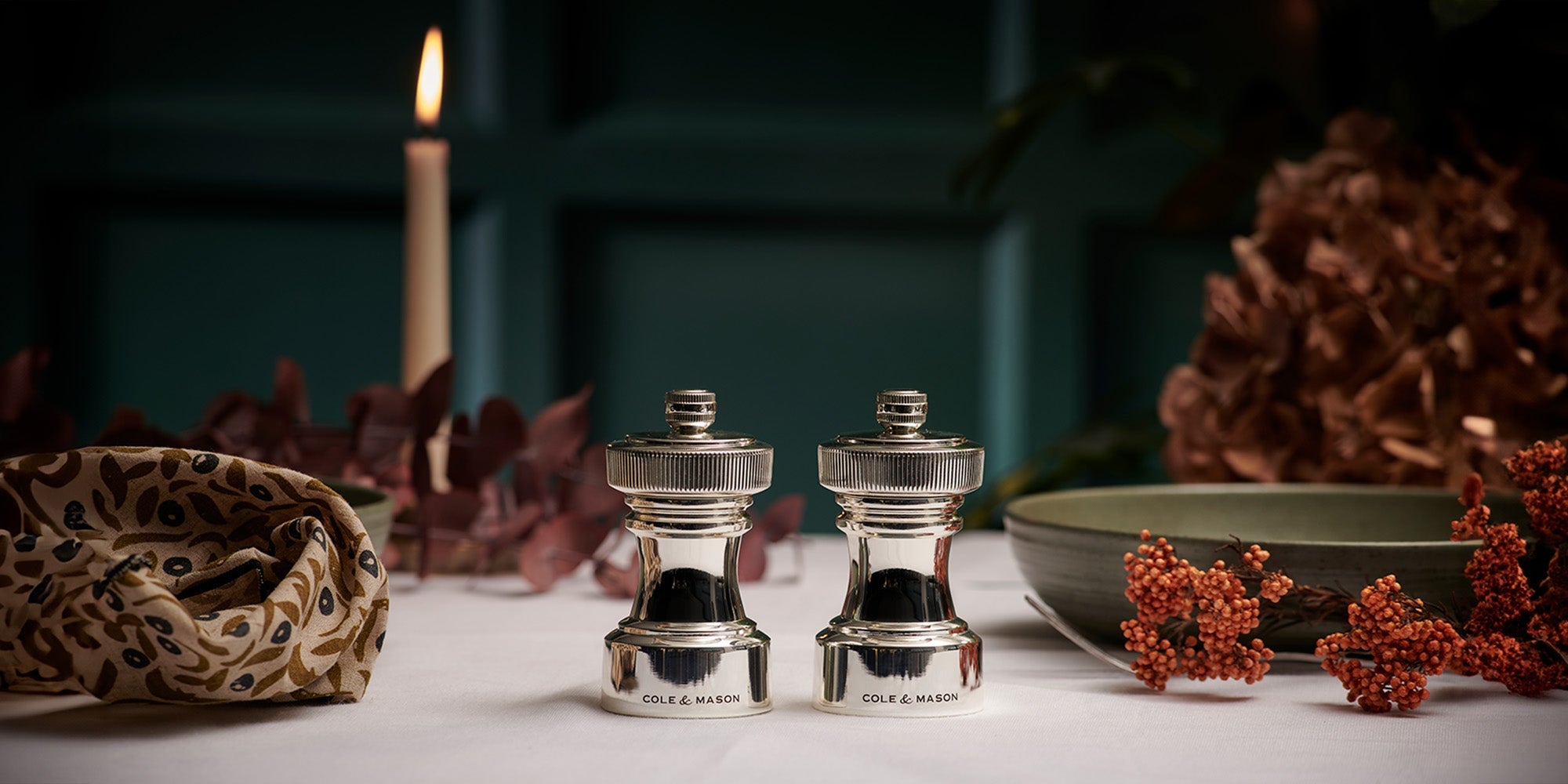 Two shiny silver salt and pepper mills sit on a white tablecloth, surrounded by dried flowers, a lit candle, a patterned napkin, and a green bowl, creating an elegant and cozy dining atmosphere.