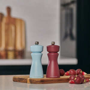 Two Cole & Mason Kenton Blue Salt & Pepper Mills, both refillable and featuring durable ceramic mechanisms, sit on a wooden board on the kitchen counter near red radishes with blurred cutting boards in the background.