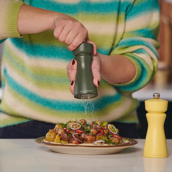 Wearing a striped sweater, someone seasons their dish with a green Cole & Mason Kenton Rosemary Salt & Pepper Mill featuring interchangeable knobs and a ceramic mechanism, while a yellow grinder sits nearby among herb- and vegetable-garnished food.