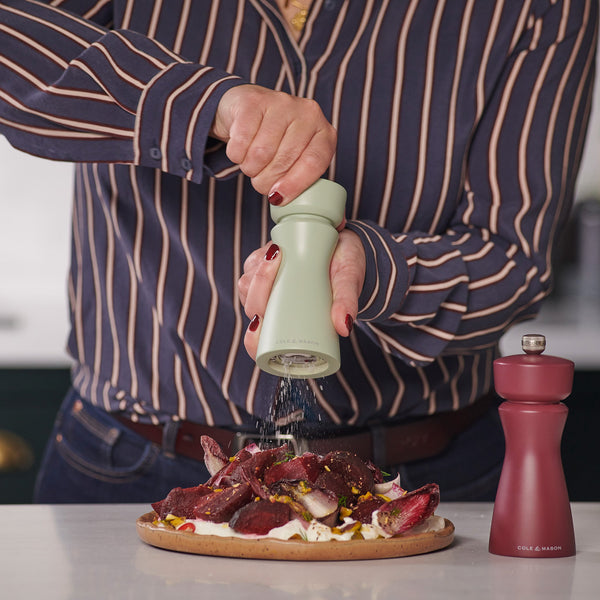 A person in a striped shirt grinds pepper onto food with the Cole & Mason Kenton Sage Salt & Pepper Mill, featuring a ceramic mechanism. A red grinder with interchangeable knobs rests on the white countertop nearby.