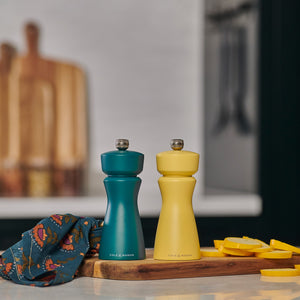 The Cole & Mason Kenton Sea Salt Blue Salt & Pepper Mill sits on a wooden board with sliced yellow squash and a patterned cloth, bringing a pop of style and featuring a high-performance ceramic mechanism for contemporary kitchens.