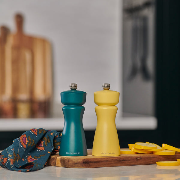 The Cole & Mason Kenton Sea Salt Blue Salt & Pepper Mill sits on a wooden board with sliced yellow squash and a patterned cloth, bringing a pop of style and featuring a high-performance ceramic mechanism for contemporary kitchens.