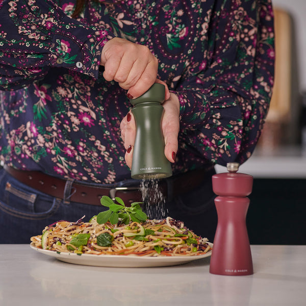Wearing a floral shirt, a person grinds black pepper onto pasta with a green Cole & Mason Kenton Rosemary Salt & Pepper Mill featuring a ceramic mechanism, while a red grinder with interchangeable knobs sits on the table.
