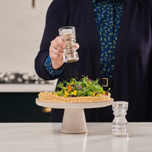 #130mm A person sprinkles pepper from a Cole & Mason grinder onto a tart with fresh greens and edible flowers on a pedestal, while the Cole & Mason London Acrylic Salt & Pepper Shaker sits on the counter nearby.