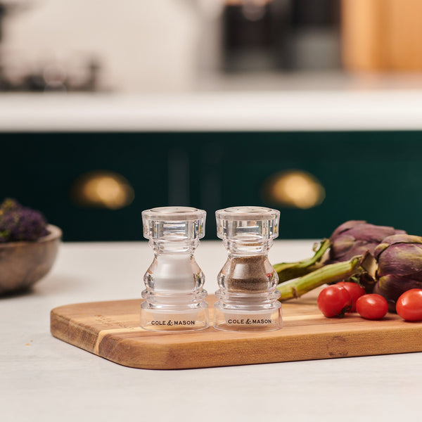 #86mm 
Two clear Cole & Mason London Acrylic Salt & Pepper Shakers sit on a wooden cutting board with tomatoes and artichokes in a kitchen featuring green cabinets and a blurred background.
