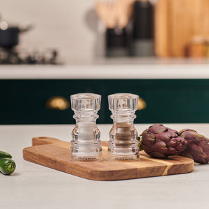 #130mm #shakerset Two transparent Cole & Mason London Acrylic Salt & Pepper Shakers sit on a wooden cutting board beside fresh artichokes on a kitchen counter, with a stove and utensils softly blurred in the background.