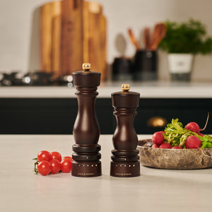 Two Cole & Mason UK London Chocolate Wood Grind Select Salt & Pepper Mills stand on a kitchen counter with cherry tomatoes and a stone bowl of radishes, offering adjustable seasoning. A blurred background shows kitchen utensils and cutting boards.