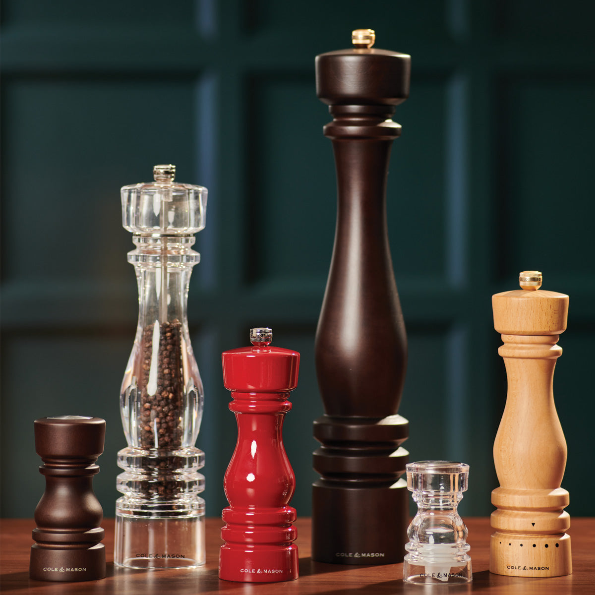 Six pepper and salt grinders of varying sizes and colors, including wood, clear acrylic, red, and black, are lined up on a wooden surface against a dark green paneled background.