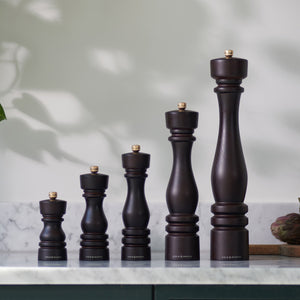 Five Cole & Mason London Chocolate Wood Salt & Pepper Mills of varying heights stand in ascending order on a marble countertop, backed by a white wall with soft natural light and plant shadows. Premium grinding technology ensures effortless use.