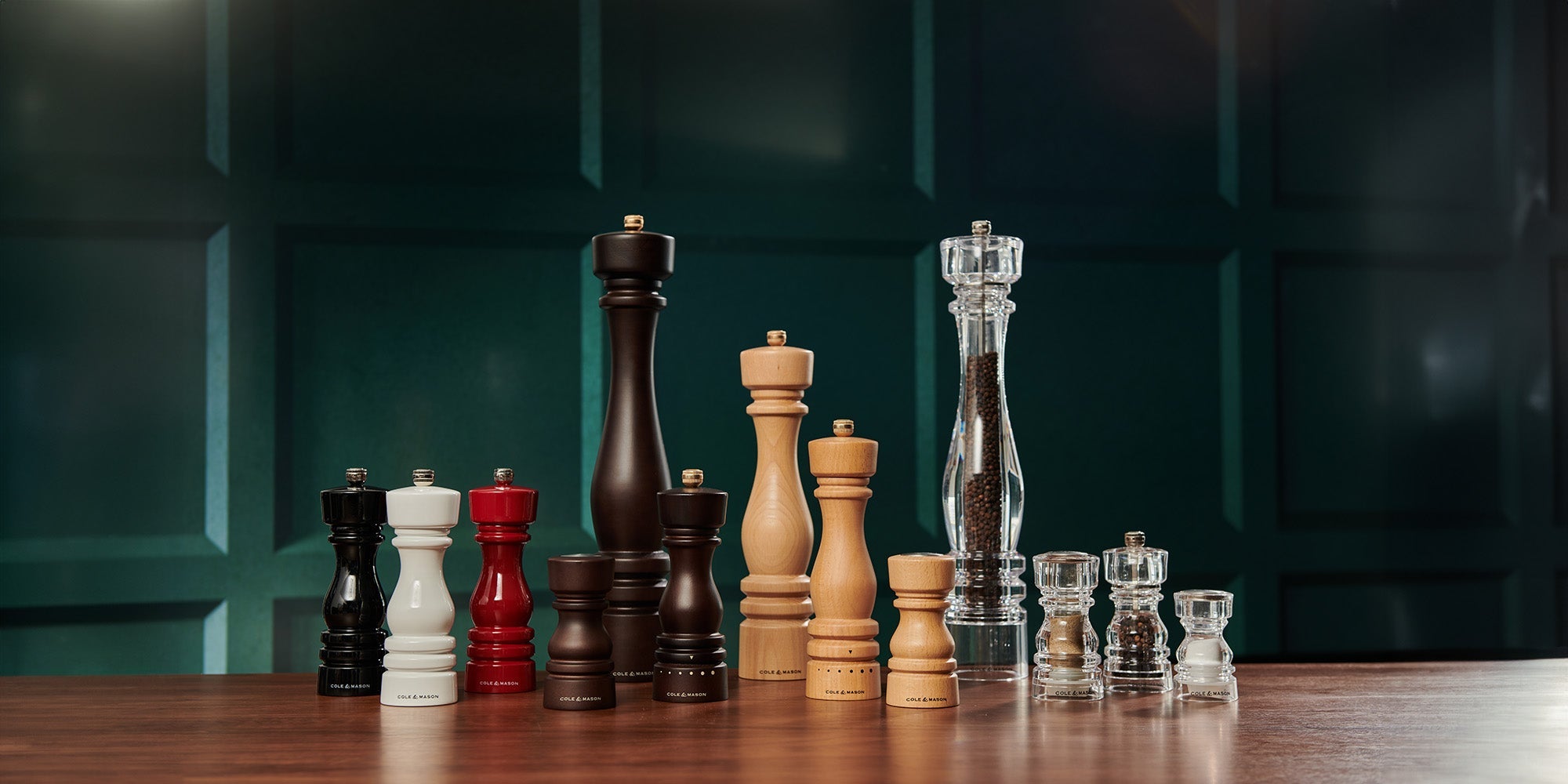 A variety of pepper and salt mills in different sizes, colors, and materials, including wood, lacquer, and acrylic, are arranged on a wooden surface against a dark green paneled background.