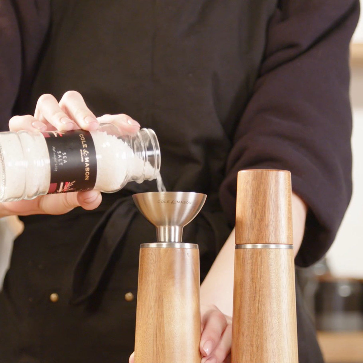 A person pours coarse sea salt from a jar into a wooden salt grinder using a metal funnel. Another wooden grinder stands nearby on the counter.