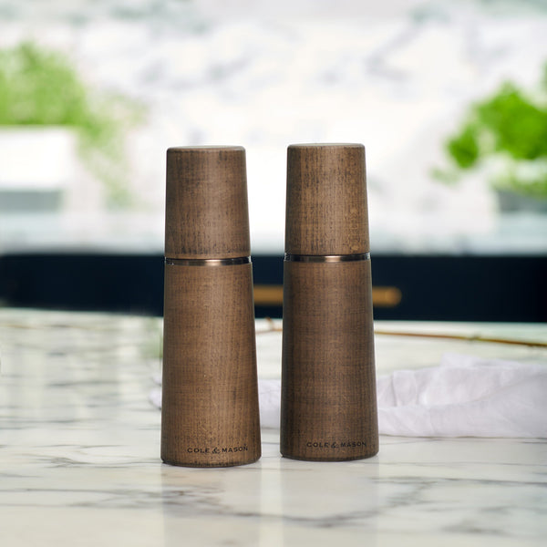 Two Cole & Mason UK Marlow Grey Beech Salt & Pepper Mills sit side by side on a white marble countertop, their sleek design complemented by blurred green plants and a white napkin in the background for a modern touch.