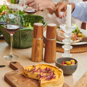 A table set for a meal features a quiche, green salad, olives, red wine glasses, Marlow Natural Acacia Salt & Pepper Mills by Cole & Mason UK with an adjustable grinder for precise seasoning, and a lit white candle in a glass holder.