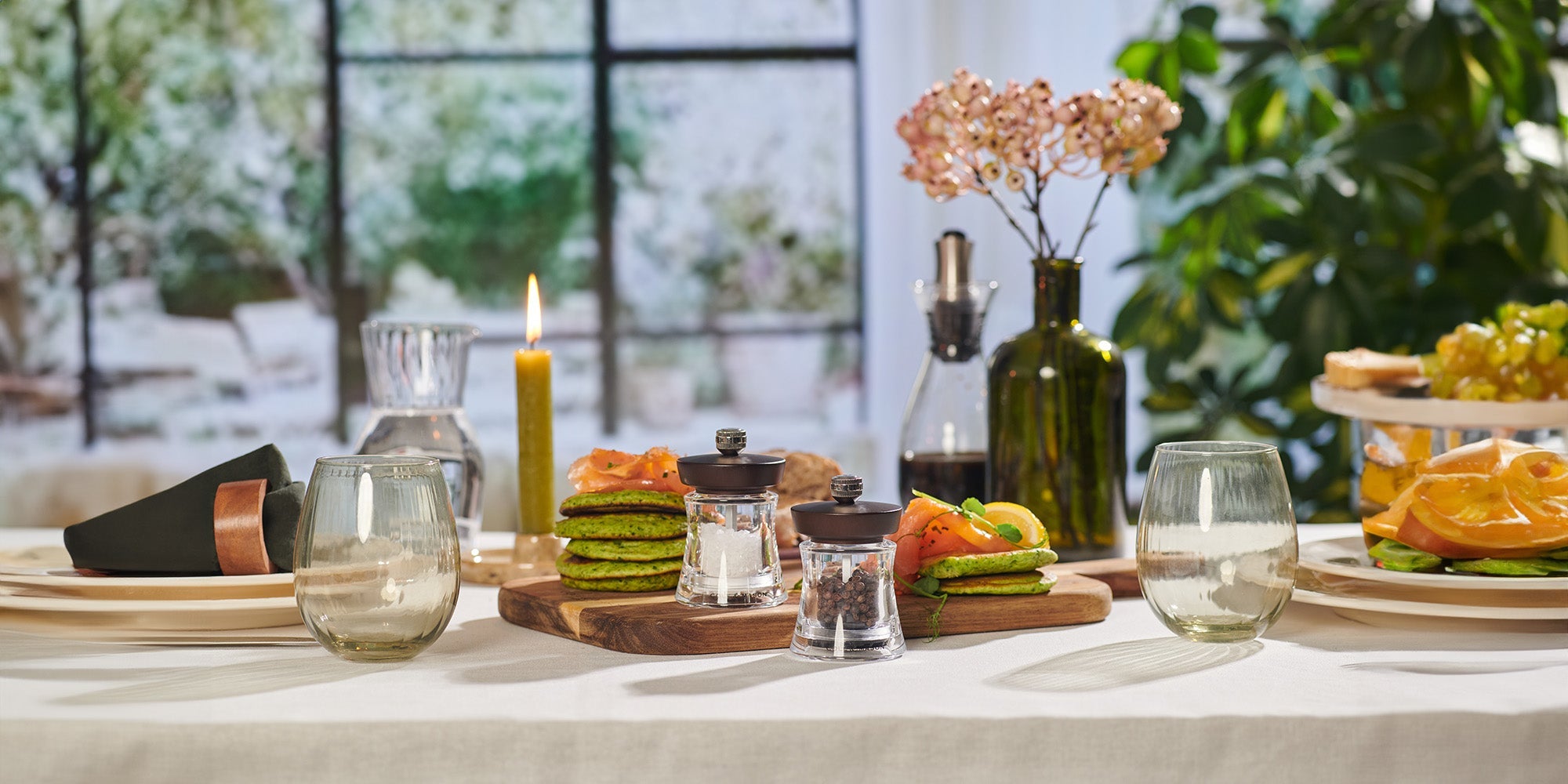 A table set for a meal with glassware, plates, bread, green pancakes topped with salmon, a lit candle, and a vase with flowers. In the background, there are plants and a large window with outdoor views.