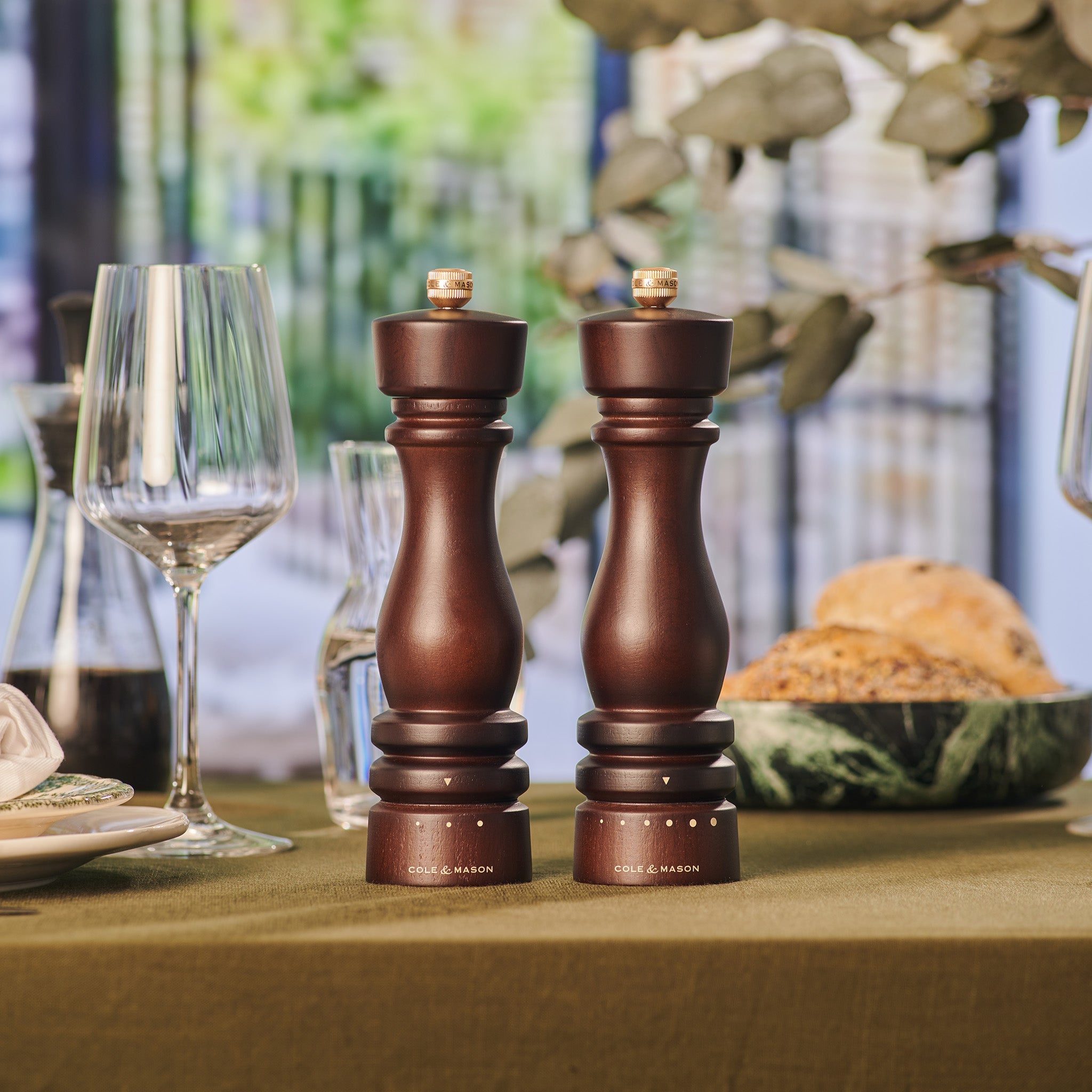 Two dark wooden pepper and salt mills stand side by side on a green tablecloth, surrounded by glassware, plates, and a bowl of bread, with blurred greenery visible in the background.