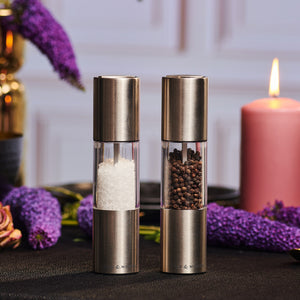 The Cole & Mason Oslo Salt & Pepper Mill Set features metallic mills with adjustable grinders, filled with white salt and black peppercorns. They sit on a dark table with glowing flowers and a lit pink candle in the background.