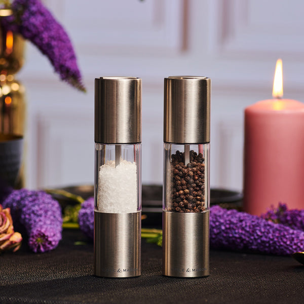 The Cole & Mason Oslo Salt & Pepper Mill Set features metallic mills with adjustable grinders, filled with white salt and black peppercorns. They sit on a dark table with glowing flowers and a lit pink candle in the background.