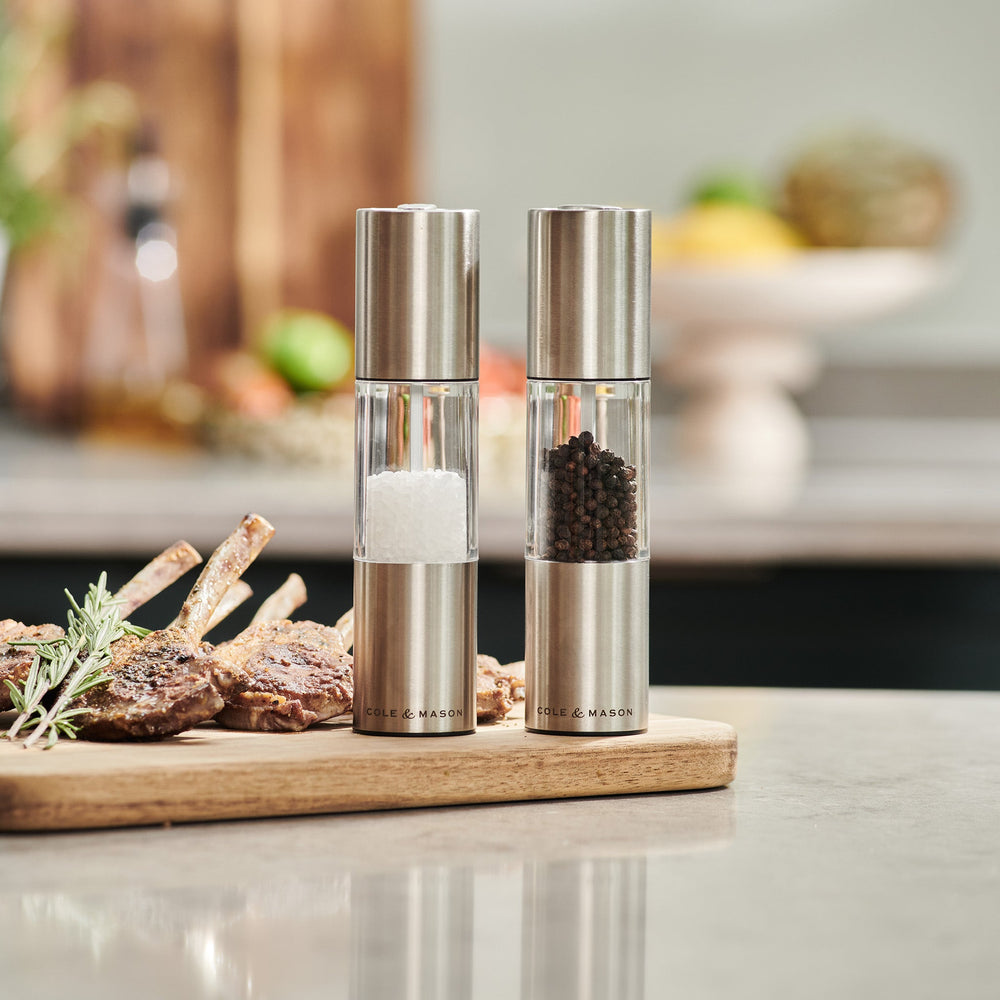 The Cole & Mason Oslo Salt & Pepper Mill Set features two upright stainless steel and glass grinders, each labeled at the base, with an adjustable mechanism for precise seasoning. Their sleek design stands out on a light background.