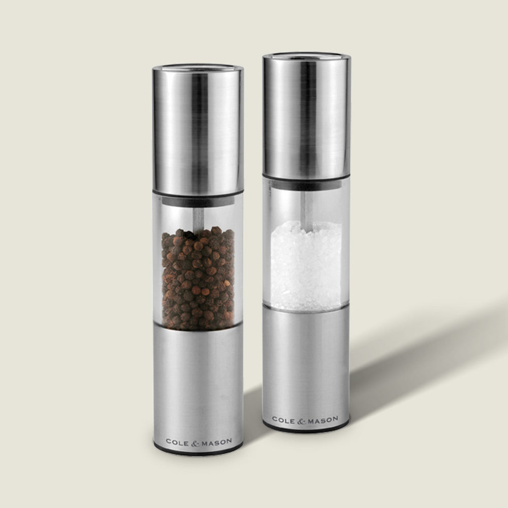The Cole & Mason Oslo Salt & Pepper Mill Set features two upright stainless steel and glass grinders, each labeled at the base, with an adjustable mechanism for precise seasoning. Their sleek design stands out on a light background.