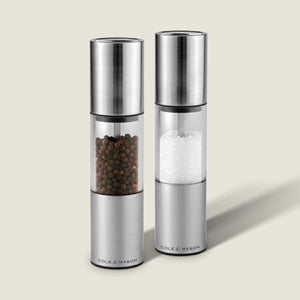 The Cole & Mason Oslo Salt & Pepper Mill Set features two upright stainless steel and glass grinders, each labeled at the base, with an adjustable mechanism for precise seasoning. Their sleek design stands out on a light background.