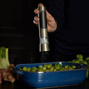 A person seasons Brussels sprouts in a blue baking dish with the Cole & Mason Richmond Electronic Salt & Pepper Mill Set, which offers adjustable grind levels. Fresh vegetables are seen on the dark counter nearby.