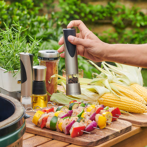 A hand seasons vegetable and chicken skewers with the Cole & Mason Richmond Electronic Salt & Pepper Mill Set on a wooden outdoor table, surrounded by fresh corn, herbs, oil, spices, and a nearby grill.