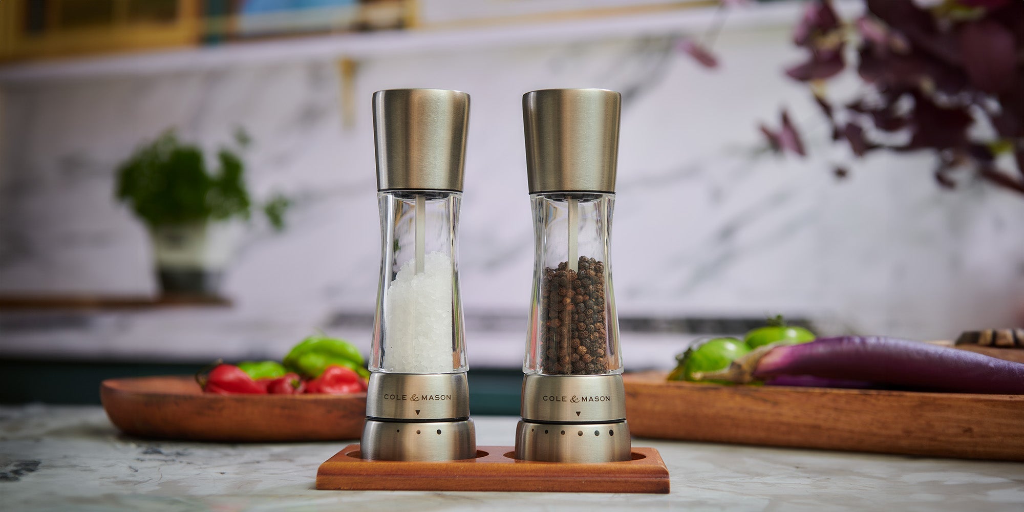 Two stainless steel and glass grinders, one filled with salt and the other with pepper, sit on a wooden tray on a marble kitchen counter. Fresh vegetables and herbs are visible in the background.