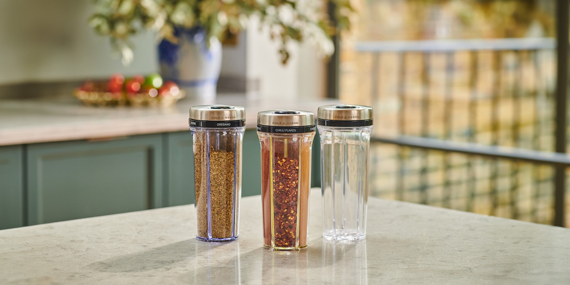Three clear spice grinders with stainless steel lids, each containing different spices, are arranged on a light kitchen countertop. A blurred vase with flowers and fruit bowl are visible in the background.