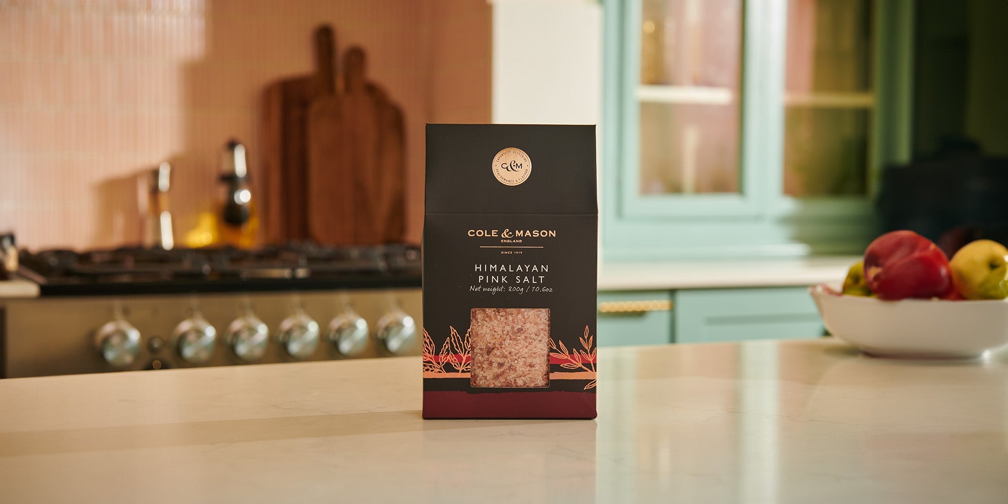 A box of Cole & Mason Himalayan Pink Salt sits on a white kitchen countertop, with a stove and wooden boards in the background and a bowl of apples on the right.
