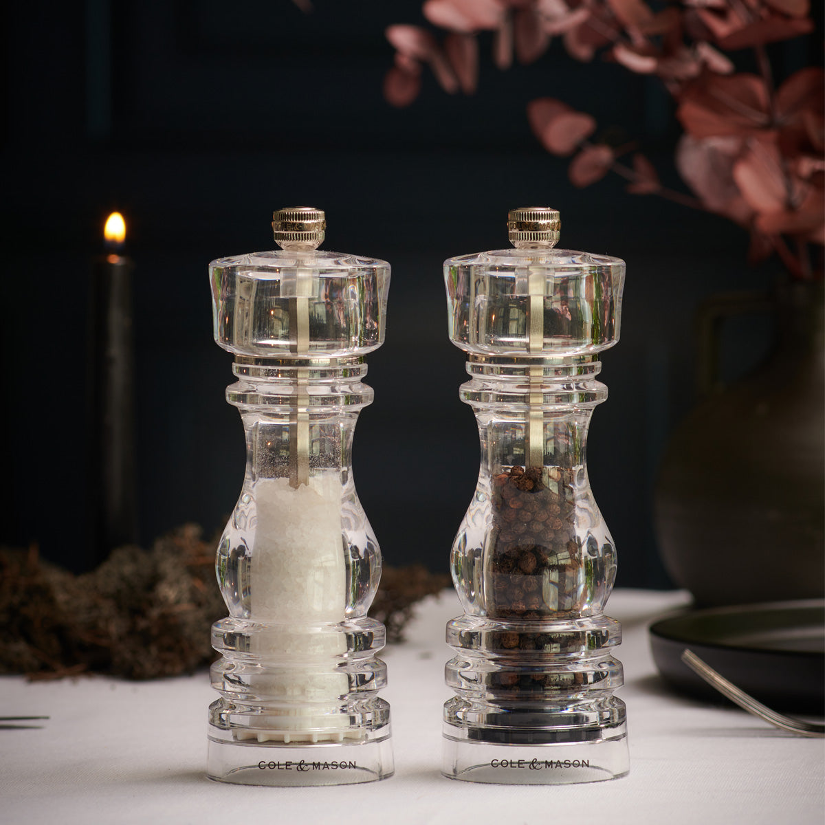 Two clear acrylic mills, one filled with white salt and the other with black peppercorns, sit on a table set for dining with dark decor, dried flowers, a candle, and plates in the background.