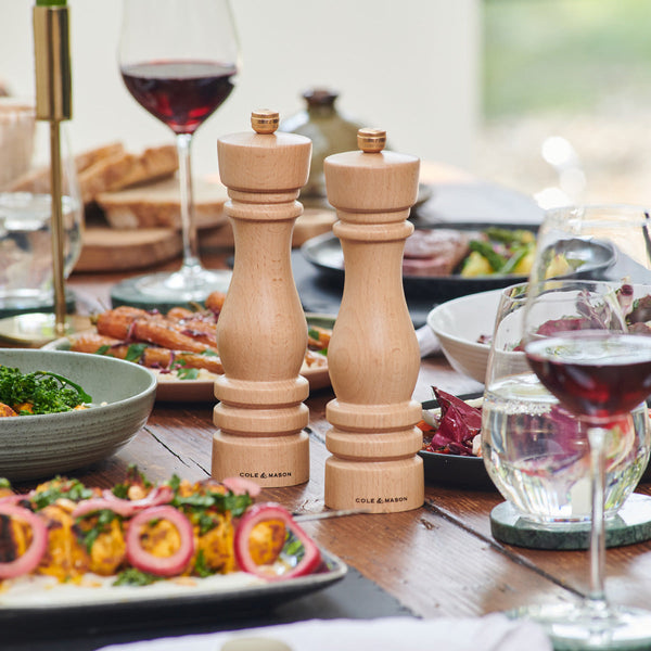 Two wooden salt and pepper grinders sit on a dining table set with plates of colorful food, glasses of red wine, and various dishes, creating a cozy, inviting meal setting.
