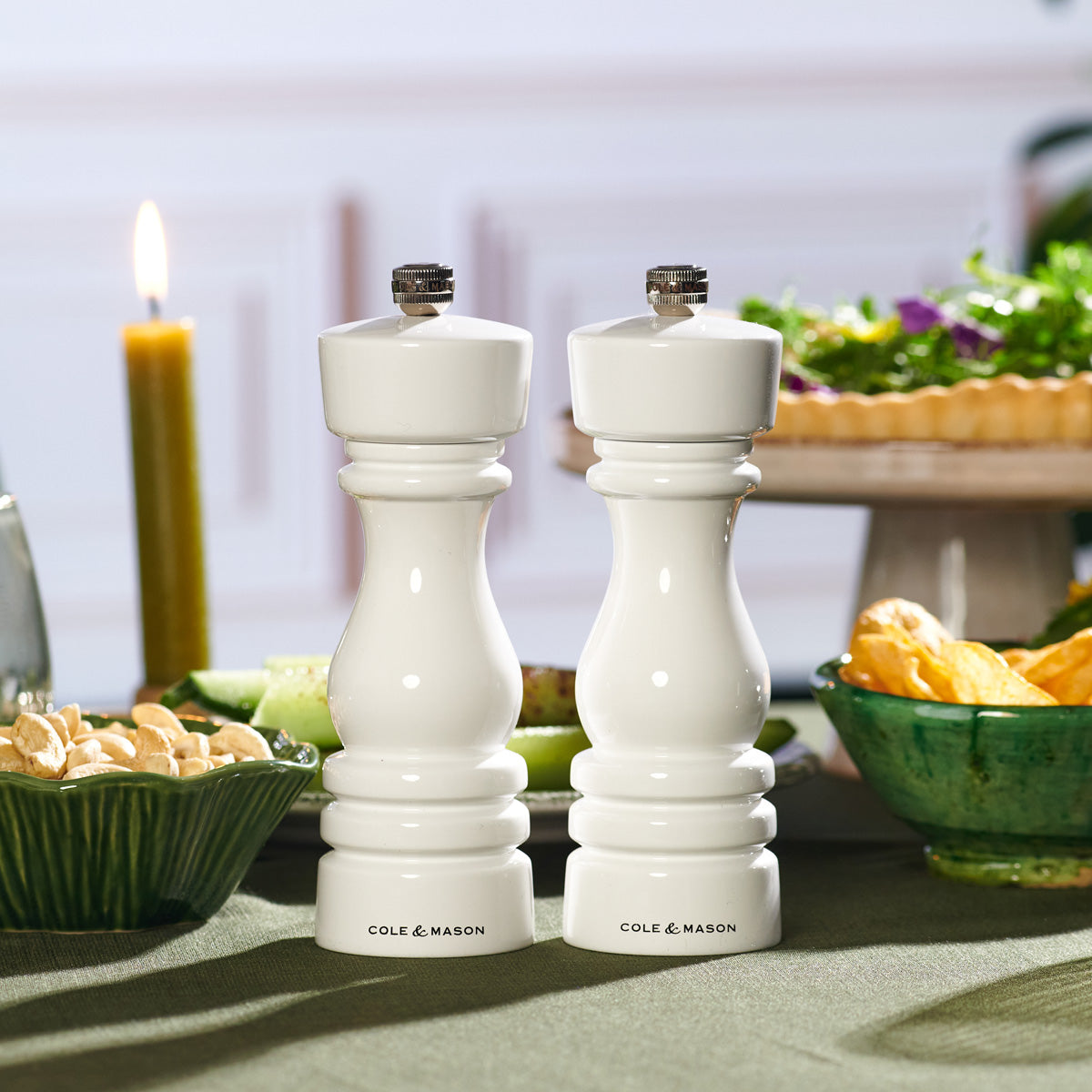 Two white Cole & Mason salt and pepper grinders are placed on a green tablecloth, surrounded by bowls of snacks, a salad, and a lit candle in the background.